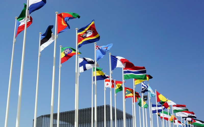 Various flags from different countries blowing in the wind against a clear blue sky