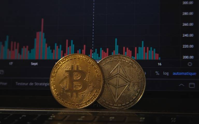 Two coins with crypto and bitcoin symbols, balancing against a laptop screen displaying a graph