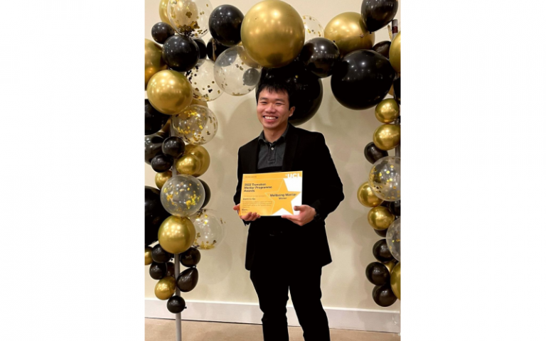Dominic Ko holding his Transition Mentorship Programme Award for Wellbeing Warrior, standing under a balloon arch