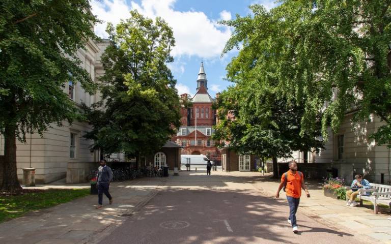 The Cruciform Building is in the distance; in the foreground of the photo is the quad, with people walking through it. There are trees along the side of the quad.