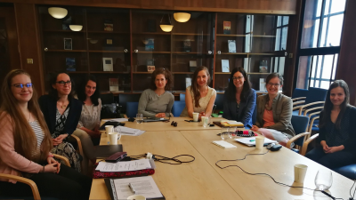 8 people part of the Feminist Judgments in Central and Eastern Europe project are sitting around a table and smiling at the camera. There are notepads, pens and laptops on the table.