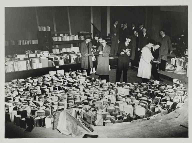 A photograph from 1941; men stand in the main library, inspecting a large number of books that stand with their pages open, drying out after being soaked in water to prevent further fire damage after the bombing of UCL's main building.