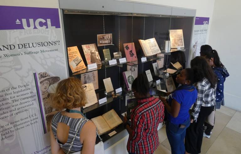 A group of young women looking at an exhibition held in UCL Main Library. The exhibition is titled 'Dangers and Delusions' and is about the women's suffrage movement. Books, photographs and magazines are on display. 
