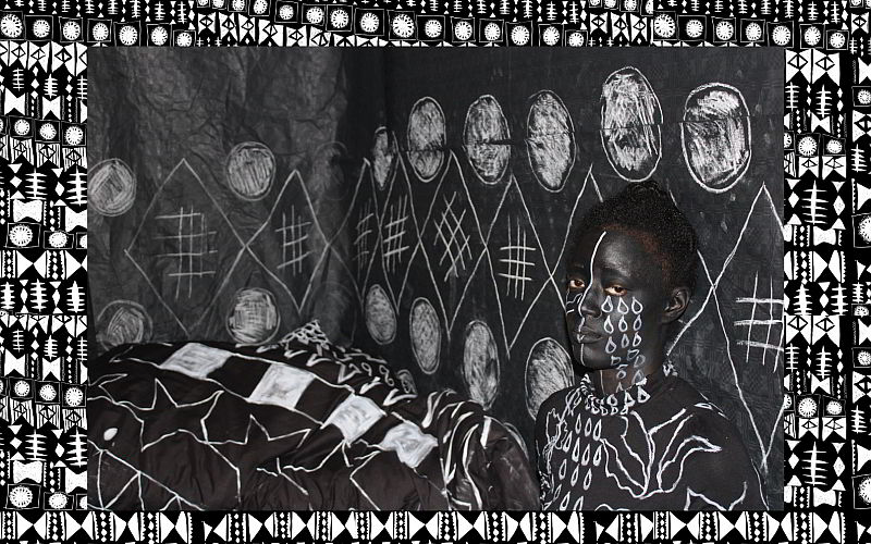 A black background with white geometric patterns on it. Superimposed over this is a photograph of a woman sitting in her bedroom. The walls, bed duvet, and woman are painted black with white geometric patterns.