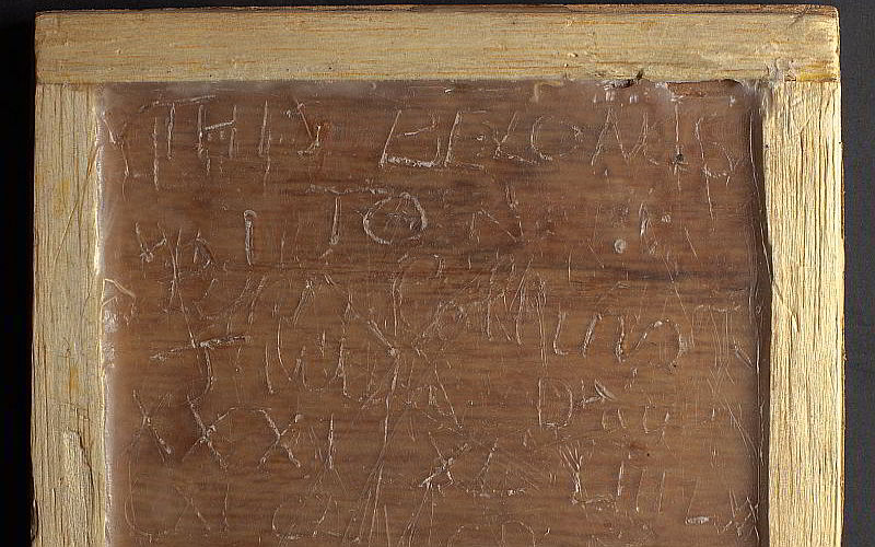 A small wooden framed slate with wax in the centre. Three different children have scratched writing into the table. They have written over each other, making the tablet difficult to read.