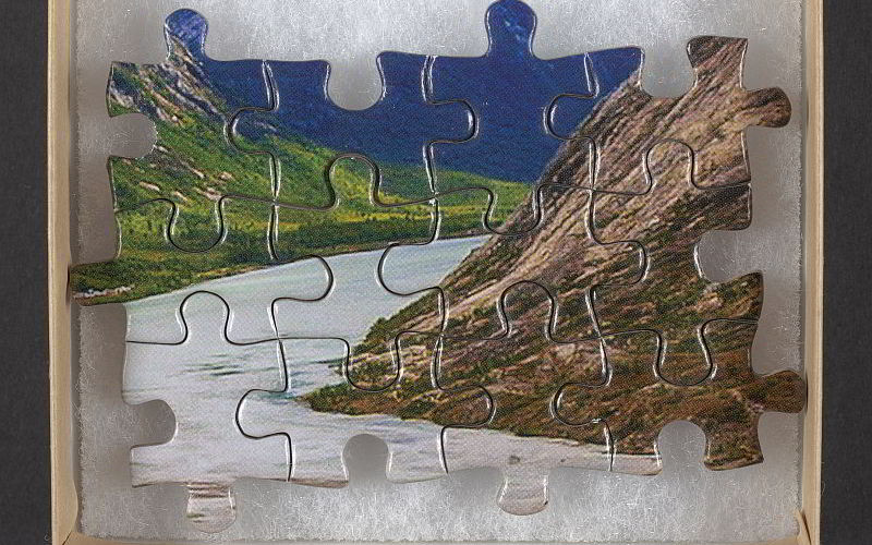 A few puzzle pieces that have been fitted together inside a small box. On top of the puzzle pieces a landscape showing a river and two hills has been painted.