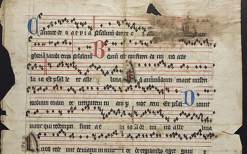 A page of parchment, cut to the shape of a book cover but with a corner section cut out. The manuscript features modal early music notation. The staff is drawn in red ink, notes in black diamond shapes and words are handwritten in black with red and blue.