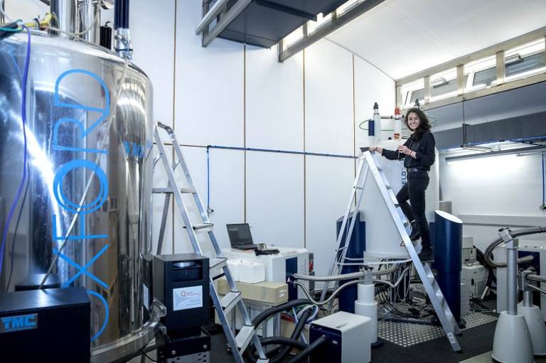 Dr Gabriella Heller on a ladder next to a a Nuclear Magnetic Resonance Spectrometer (NMR) machine.