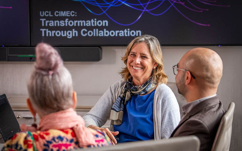 Partners discuss with CIMEC team members around a table in a conference room in Bloomsbury. UCL CIMEC branding on screens at the rear.