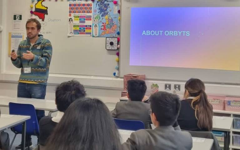 A researcher standing at the front of a classroom with the backs of heads. On the board it says “About Orbyts”