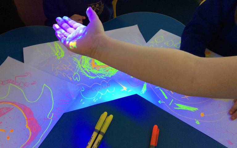 Picture of a child’s arm with glowing colours under a UV light, in the background you can see paper with felt tip drawings