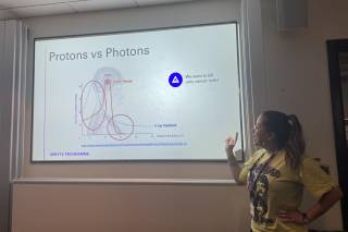 A female researcher standing in front of a white board that says “Protons vs Photons”