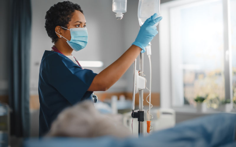 A healthcare worker in a hospital ward, checking the status of an intravenous drip bag.