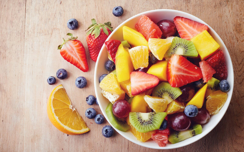 View from above of a bowl of fruit salad, including chopped strawberries, kiwi and various types of berries, with some spilling over onto a kitchen worktop