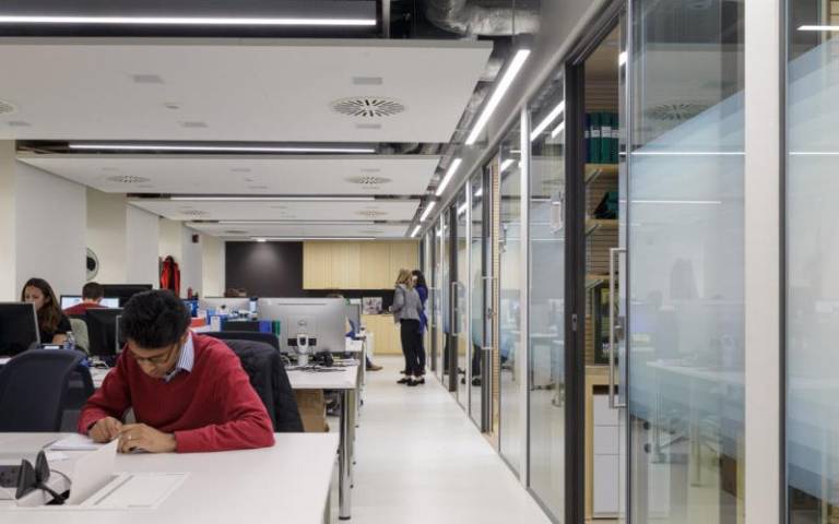 UCL staff working in open-plan desk space with glass screens and doors to laboratories on the right 