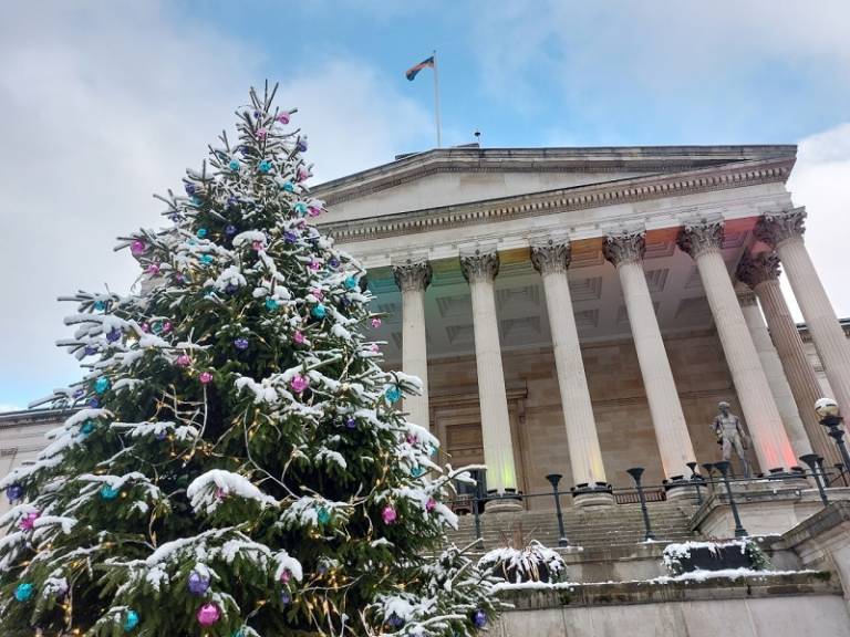 UCL's Christmas tree in the Quad, December 2023. The tree is in the foreground, decorated with baubles. The Quad is in the background, lit up in rainbow colours. It is day time and there is a covering of snow over everything.
