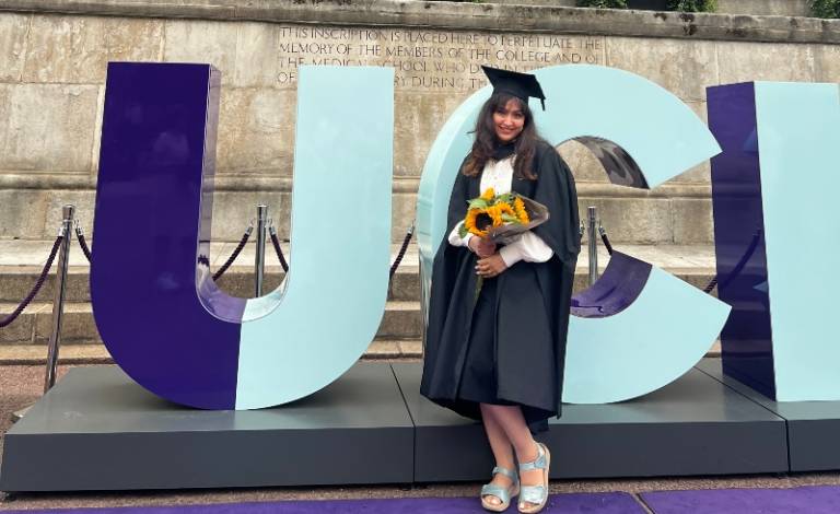 Arifa standing in front of the big UCL signage. She is wearing her graduation gown and holding a sunflower bouquet.