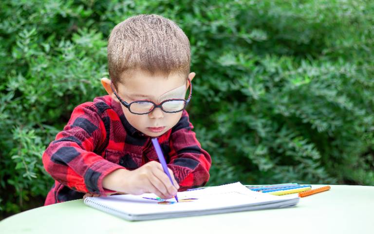 A little boy draws in the garden (outdoors). He wears glasses and an eye patch (occluder) to prevent amblyopia.