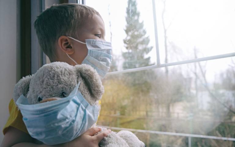 A child wearing a mask looks out a window holding a stuffed bear