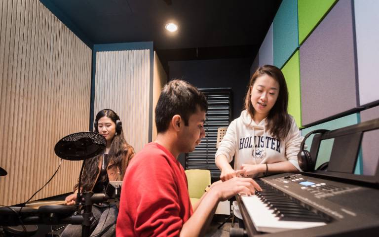 Two UCL students sit at an electronic drum kit and keyboard with a third UCL student talking to them