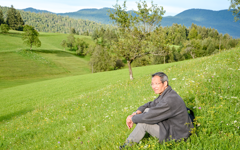 Professor Dennis Chan, sitting in a grassy meadow with trees and wooded hills in the distance
