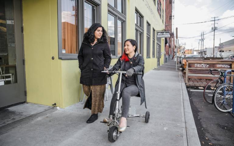 A Latinx (invisibly) disabled woman talking and walking alongside her friend, an Asian disabled genderfluid person wearing compression gloves and driving a lightweight electric mobility scooter.