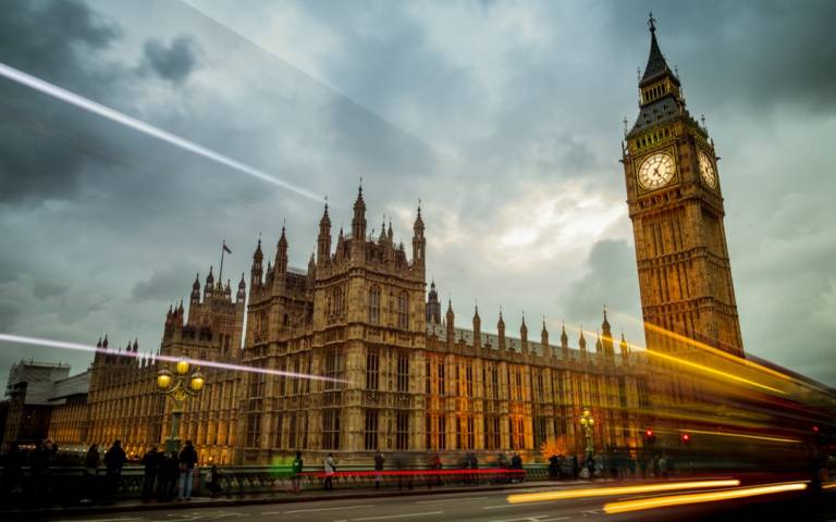 Big Ben and the Houses of Parliament in London, England. The building is lit up and set against a dramatic sky with cars and busses zooming by on Waterloo Bridge.