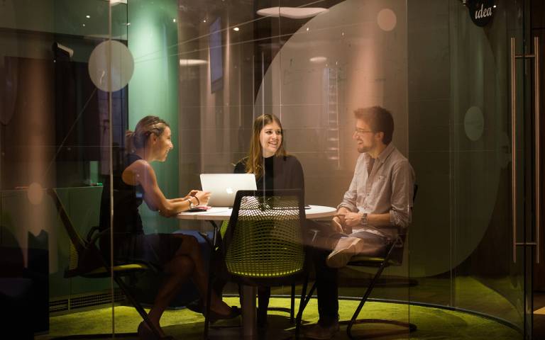 Three people sit around a table smiling and talking in a meeting room at IDEALondon
