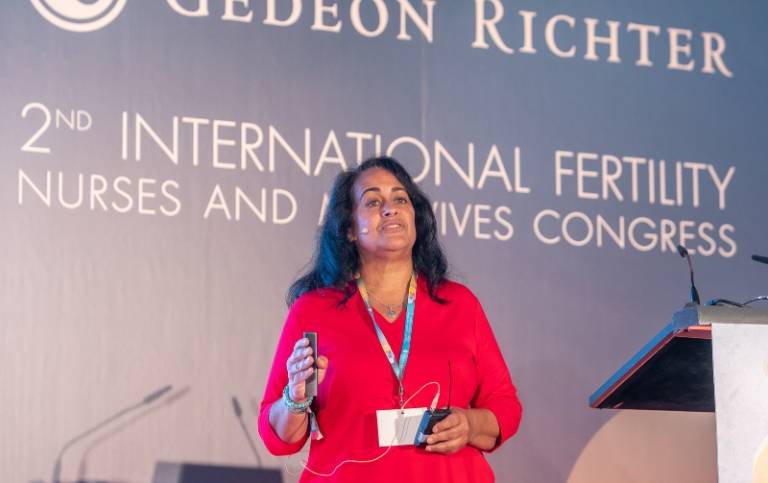 A woman wearing a red dress and microphone, giving a speech. There is a blue banner behind her that says '2nd International Fertility Nurses.'