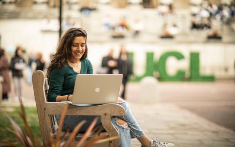 A student sits, smiling, working on her laptop outdoors in the UCL Quad