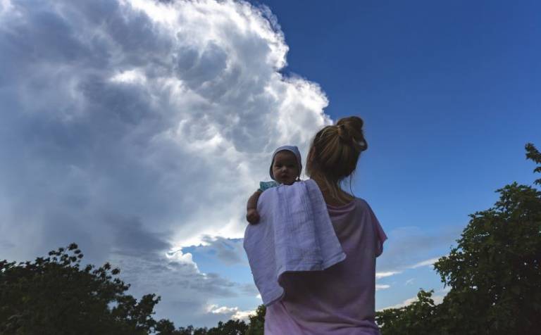 Mother holding her baby child daughter in arms looking toward the storm cumulonimbus cloud above them, baby looking at camera with hat.