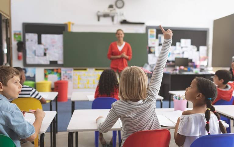 Image shows a classroom with a child raising her hand