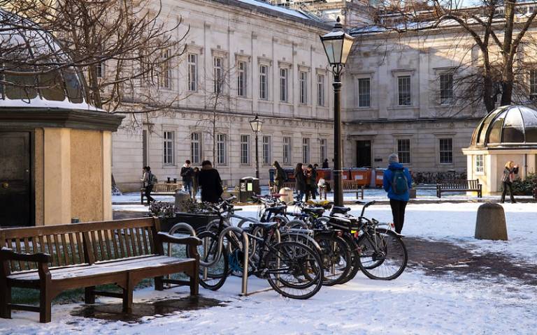 An image of UCL's Quad during snowy weather, looking towards the South Cloisters with snow on the ground, benches and observatories, parked bicycles in the foreground and people walking around dressed in warm winter clothing