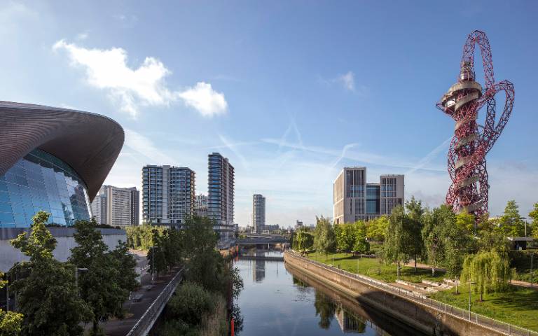 View from a distance of UCL East campus on the Queen Elizabeth Olympic Park in sunshine, showing One Pool Street just beyond the London Aquatics Centre on the left and, across the river, Marshgate just beyond the ArcellorMittal Orbit.