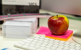 A wireless keyboard, an apple on top of two sticky note pads, and a UCL branded card holder