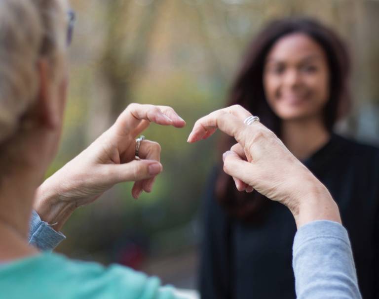 Two women using British Sign Language, with one signing 'Research'. Focus on hands, background blurred.