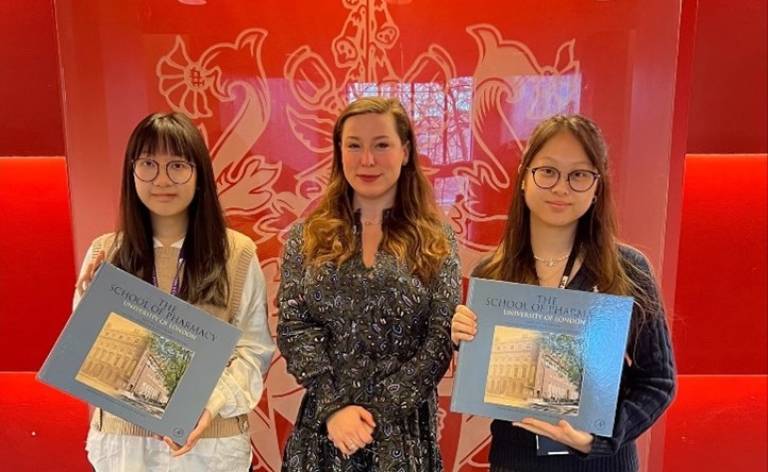 Oksana Pyzik with Stella Fu and Dorothea Tang. They are standing in front of a red wall that has a glass panel with the School of Pharmacy shield. They are holding books.