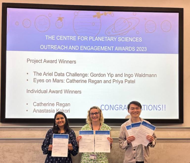 Priya Patel, Catherine Regan and Gordon Yip holding their Outreach Award certificates in front of a screen which announces their awards