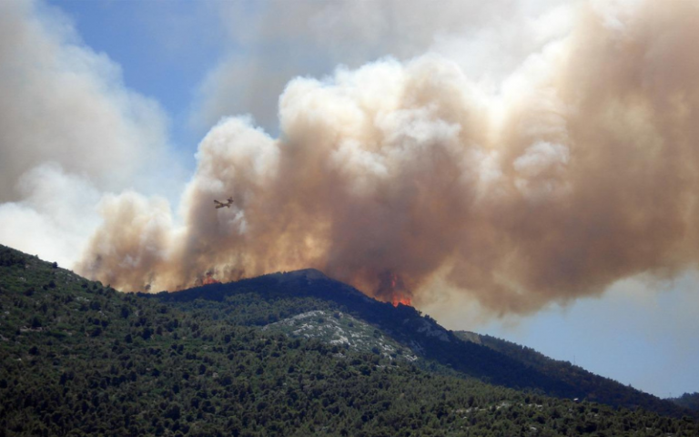A tree covered mountain is in the foreground. In the background you can see smoke and some flames rising from a mountain behind.
