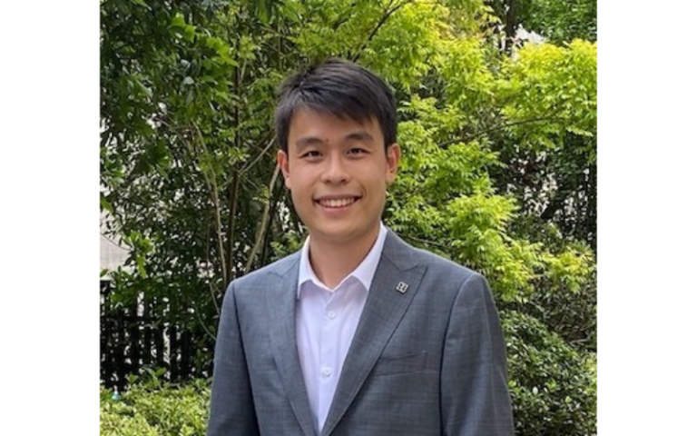 Zihan wears a grey suit and white shirt with no tie. He is standing in a park and smiling to the camera