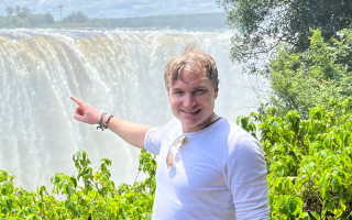 Dr Seiferling stands in front of Victoria Falls and points to towards the waterfall