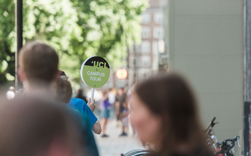 一名学生大使举着一块绿色的标牌，上面写着“伦敦大学学院校园之旅”。