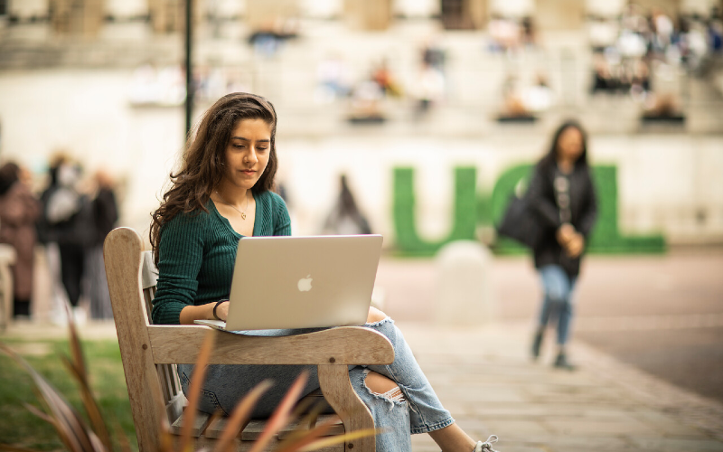 A UCL student sits on a bench in the UCL front quad and uses a laptop.