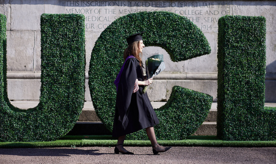 UCL graduate walks past big green "UCL" letters.