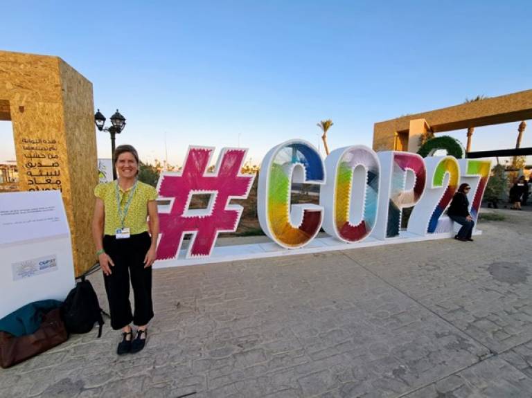 Kate stands in front of a vibrant sign which says COP27