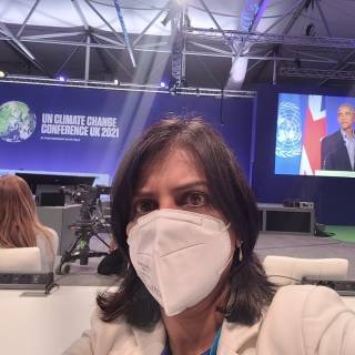 Priti stands infront of blue sign saying COP26 2021 with barack obama on a screen in the background