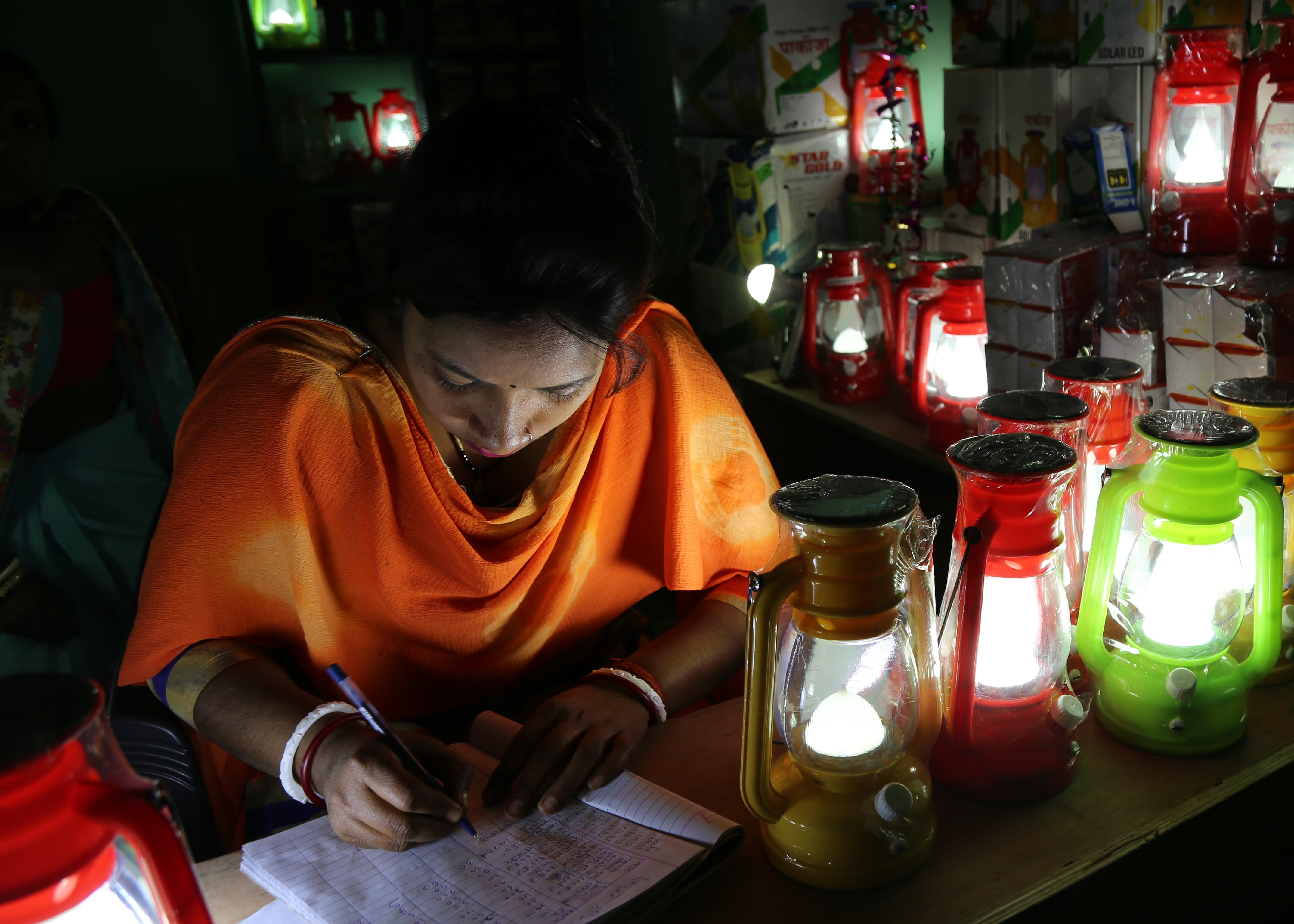 a_woman_completing_paperwork_by_the_light_of_solar_powered_lamps_in_a_village_shop_bihar.