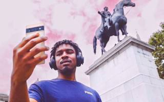 A man wearing headphones holding a smartphone up in front of a black statue of a seated rider on a horse on a white stone plinth