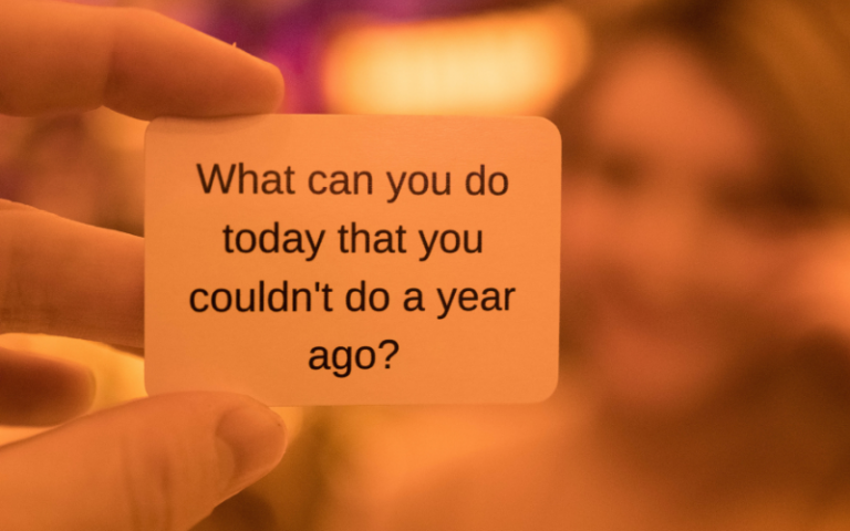 A hand holding a piece of paper with text 'what can you do today that you couldn't do a year ago?'