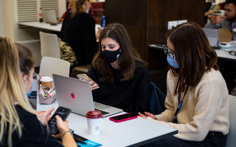 A group of young women wearing face masks sit around a table with their laptops